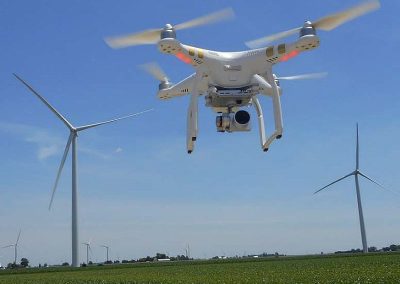 A white quadcopter drone hovers over a green field with wind turbines in the background under a clear blue sky.