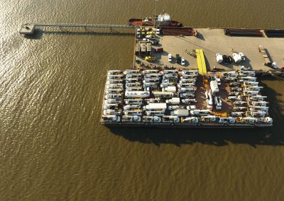 Aerial view of a dock with a barge carrying numerous white utility trucks, moored beside a pier extending into brownish water. Several vehicles and equipment are on the concrete dock.