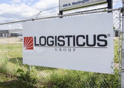 A white sign attached to a chain-link fence displays the LOGISTICUS Group logo in black letters with a red geometric design on the left side. Grass and a street address sign are visible in the background.