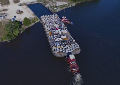 Aerial view of a large barge loaded with vehicles and equipment being pushed by tugboats on a river near a shoreline with a dock and trees.