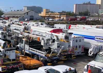 A large fleet of utility trucks and vans, including bucket trucks and vehicles marked Penske Truck Rental, is parked closely together in a lot near industrial buildings, with tall city buildings visible in the background.
