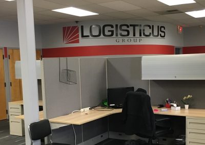 An office space with cubicles, desk chairs, computers, and office supplies. LOGISTICUS GROUP is displayed in large letters on the back wall above a red stripe. Light wood doors are closed in the background.