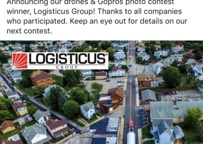 A large wind turbine blade is transported on a truck along a suburban street lined with houses, as seen from above. The image is part of a contest announcement by the Specialized Carriers & Rigging Association.