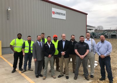 A group of eleven men stand in front of a gray industrial building with a Logisticus sign, some in business attire and others in safety vests and work clothes, posing for a group photo on a gravel lot.