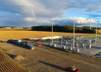 A wind farm construction site in a rural field with wind turbine parts, vehicles, and equipment. Several turbines are standing; a rainbow appears in the partly cloudy sky above the landscape.