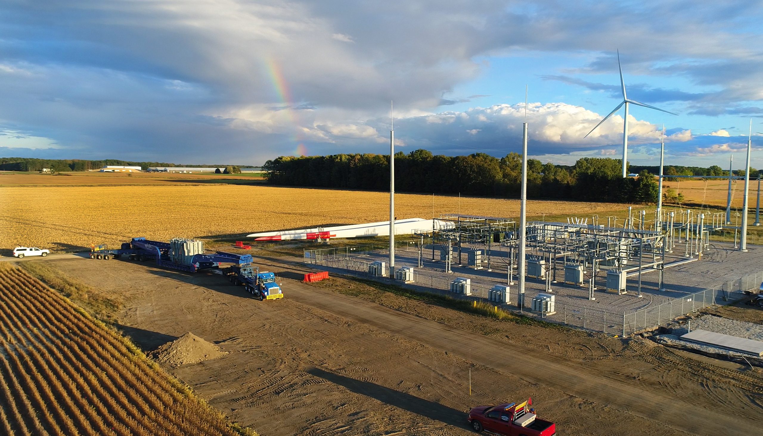 A wind farm construction site in a rural field with wind turbine parts, vehicles, and equipment. Several turbines are standing; a rainbow appears in the partly cloudy sky above the landscape.