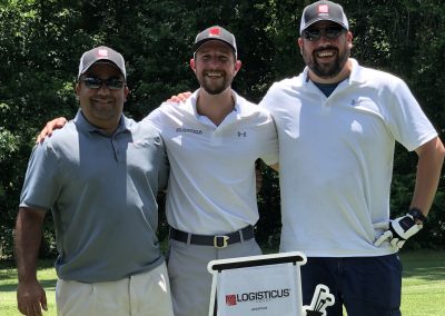 Three men stand outdoors on a golf course, smiling at the camera. They are wearing golf attire, black caps, and sunglasses. There’s a LOGISTICUS sign in front of them, with green trees in the background.