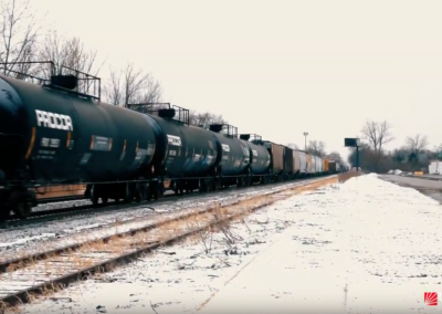 A long freight train with black tanker cars travels down snowy tracks on a cold, overcast day. Leafless trees line the background, and a few rail-side buildings are visible at the right.