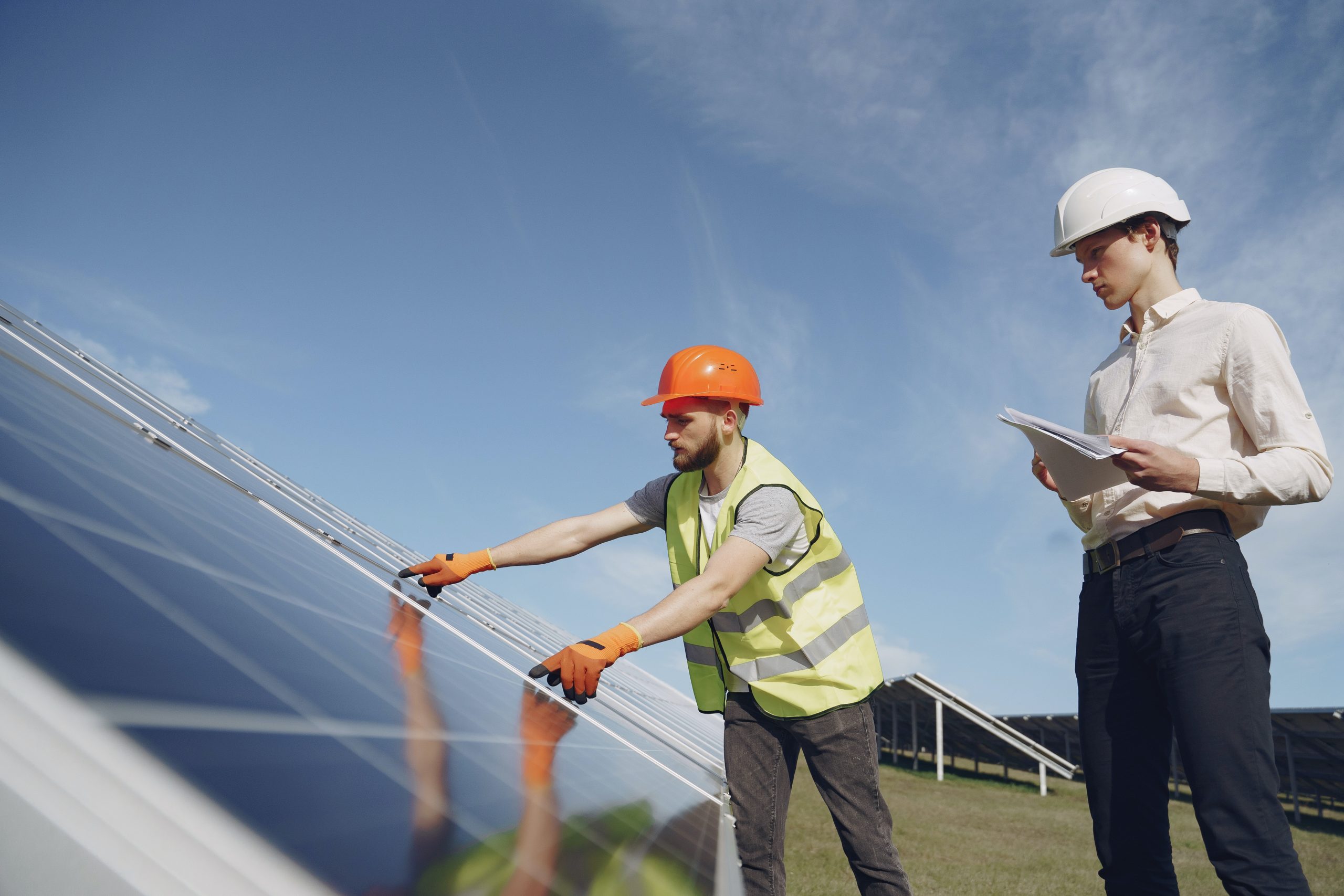 Solar Farm 1 Two workers in safety gear inspect solar panels outdoors; one adjusts a panel while the other holds documents, both wearing helmets and vests under a clear sky.