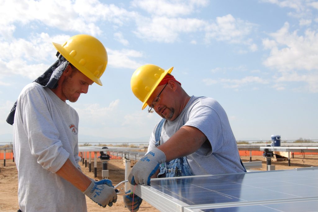 Two men wearing yellow hard hats and gloves work together installing solar panels at an outdoor construction site under a partly cloudy sky.