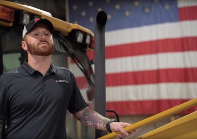 A man with tattoos wearing a black polo shirt and cap stands in front of construction equipment, holding a yellow railing. A large American flag hangs in the background.