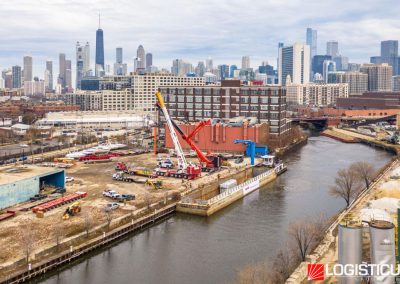 An aerial view of a construction site by a river in Chicago, with cranes, trucks, and workers visible. The city skyline, including skyscrapers, is prominent in the background. The LOGISTICUS GROUP logo is in the bottom right corner.