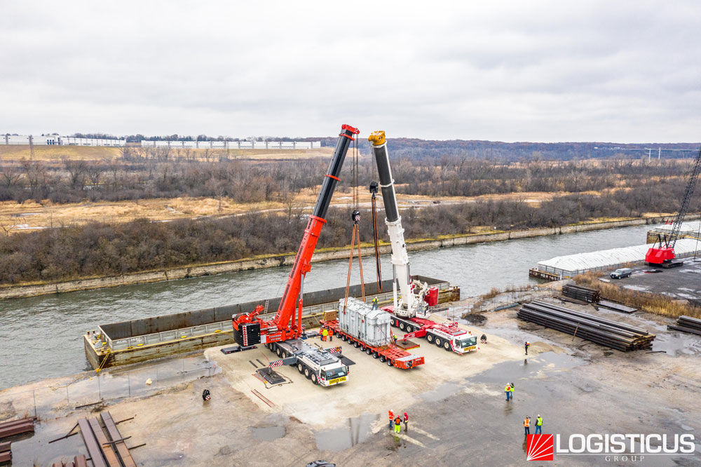 Two large cranes lift heavy equipment from a barge at a riverside industrial site, while workers in safety vests supervise. Stacks of materials and vehicles are nearby. LOGISTICUS GROUP logo is in the bottom right corner.