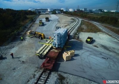 A large industrial transformer is being loaded onto a flatbed railcar using forklifts and heavy machinery. Workers in safety gear are assisting on a gravel area near train tracks and storage tanks. LOGISTICUS logo is visible.