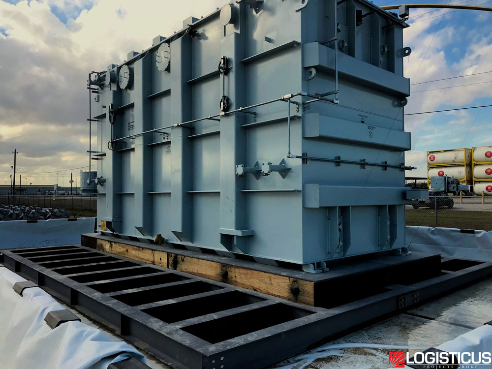 A large industrial transformer sits on a flat platform outdoors, surrounded by utility poles and a chain-link fence. The sky is partly cloudy, and the LOGISTICUS logo appears in the lower right corner.