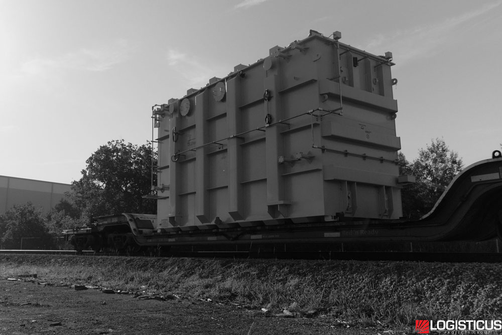 A large industrial container is secured on a flatbed train car, traveling along railroad tracks in an outdoor setting; trees and a building are visible in the background. The image is in black and white.
