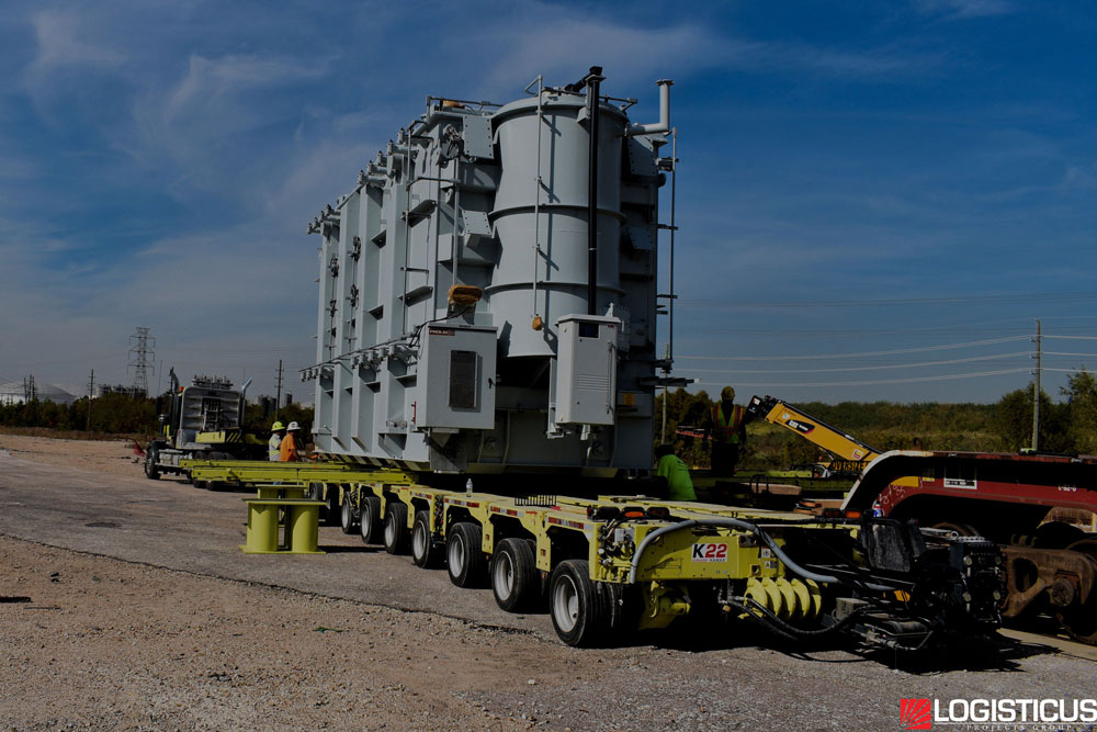 A large industrial transformer is loaded onto a multi-axle flatbed trailer for transport, with several workers nearby and power lines visible in the background. The image is branded with the LOGISTICUS logo.