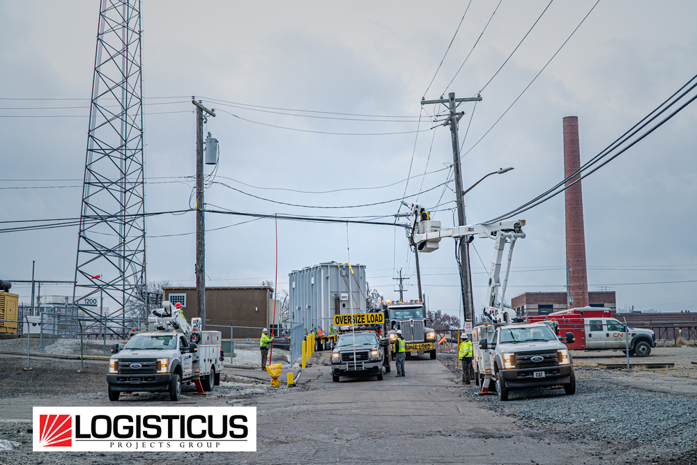 Utility workers in safety gear use bucket trucks to manage power lines as an OVER SIZE LOAD vehicle transports large equipment down an industrial street. LOGISTICUS Projects Group logo is visible in the lower left corner.