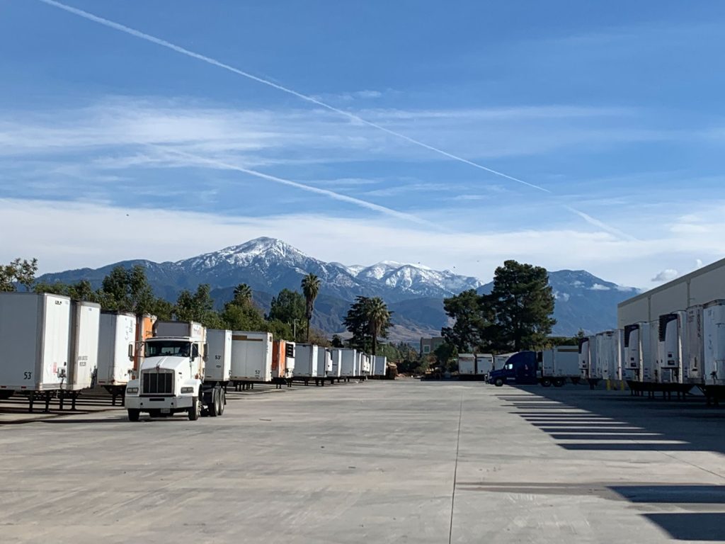 A large parking lot with rows of parked white semi-trailers and a white truck, set against a backdrop of snow-capped mountains under a blue sky with thin clouds and contrails.