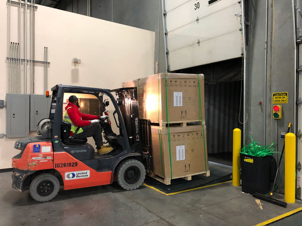 A worker in a red jacket operates a forklift to load large cardboard boxes, wrapped in green plastic, into a truck at an indoor loading dock. Safety caution signs and yellow barriers are visible nearby.