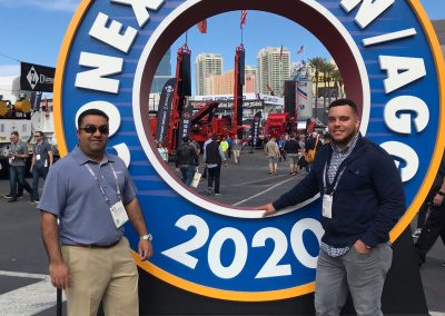 Two men stand posing by a large circular CONEXPO CON/AGG 2020 sign at an outdoor event with construction equipment and city buildings in the background.