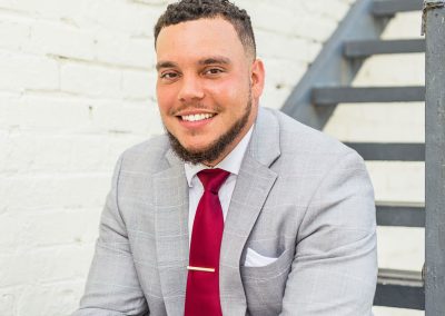 A man with short curly hair and a beard, wearing a light gray suit, white shirt, and red tie, smiles while sitting next to a metal staircase against a white brick wall.