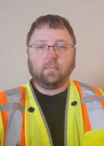 A man with glasses, short brown hair, and a beard wears a black shirt under a yellow safety vest with orange and silver reflective stripes, standing against a plain light-colored background.