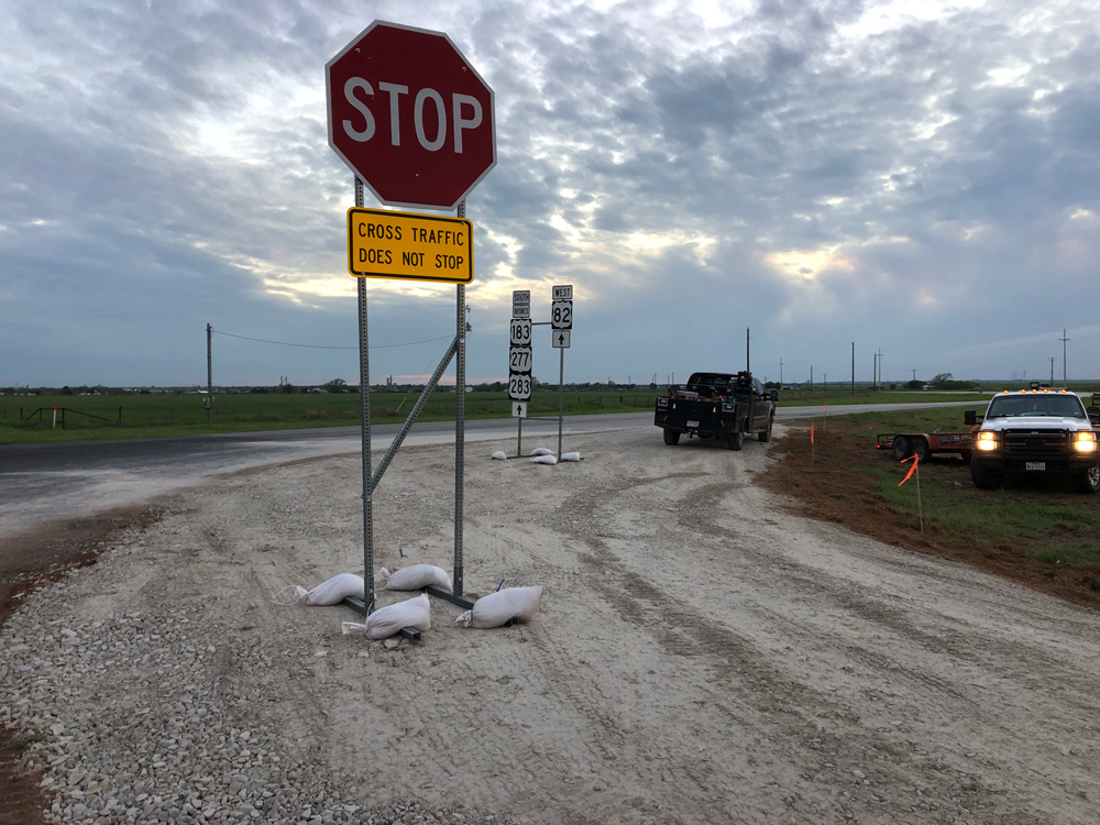 A stop sign with a yellow Cross Traffic Does Not Stop sign stands on a gravel road; sandbags anchor the sign. Two vehicles are parked nearby with cloudy skies overhead and open fields in the background.