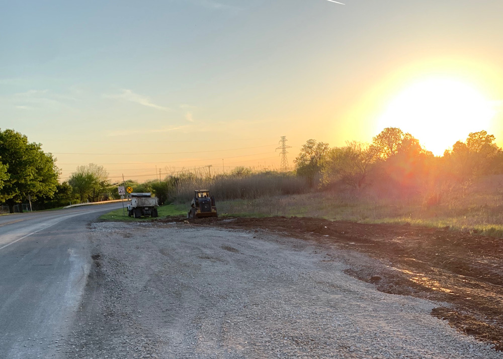 A rural roadside construction site at sunset, with a dump truck and a small bulldozer parked on gravel beside a road. Trees and power lines are visible in the background under a clear sky.