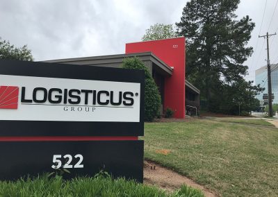 A black and white Logisticus Group sign with a red logo stands on a lawn in front of a modern building marked 522, with trees and a cloudy sky in the background.