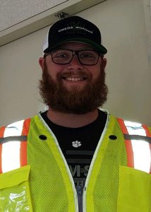 A person with a beard and glasses smiles, wearing a black cap, black shirt, and a high-visibility yellow safety vest with reflective stripes, standing indoors.