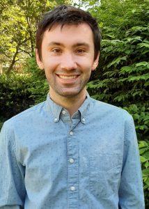 A man with short dark hair, wearing a light blue button-up shirt, smiles while standing outdoors in front of lush green bushes and trees.