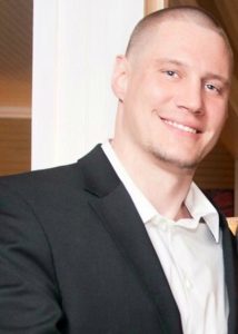 A man with short hair, wearing a black suit jacket over a white dress shirt, stands indoors and smiles at the camera.