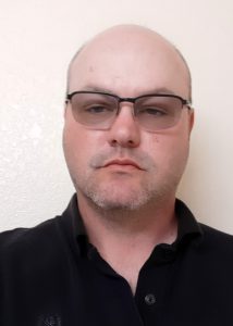 A man with short hair, wearing glasses and a black collared shirt, stands against a plain, light-colored wall, looking directly at the camera with a neutral expression.