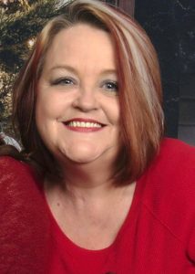 A smiling woman with short, straight, reddish-brown hair with light highlights, wearing a red top, poses indoors with a blurred background.