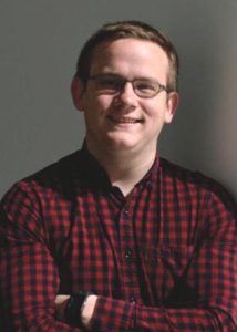 A person wearing glasses and a red and black checkered shirt is smiling with arms crossed, standing against a plain gray background.