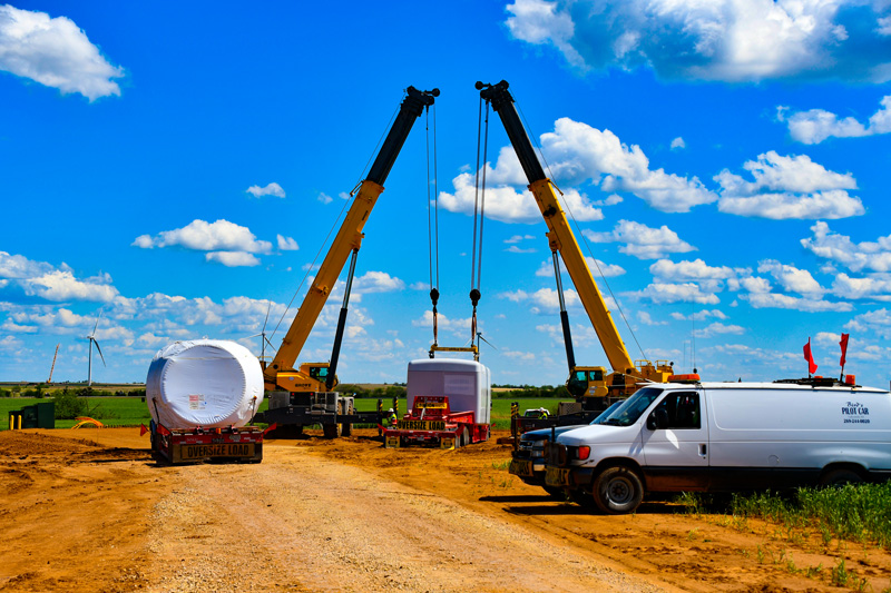 Two large cranes lift equipment on a construction site with a dirt road, a white van, and a flatbed truck under a bright blue sky with scattered clouds. Fields and wind turbines are visible in the background.