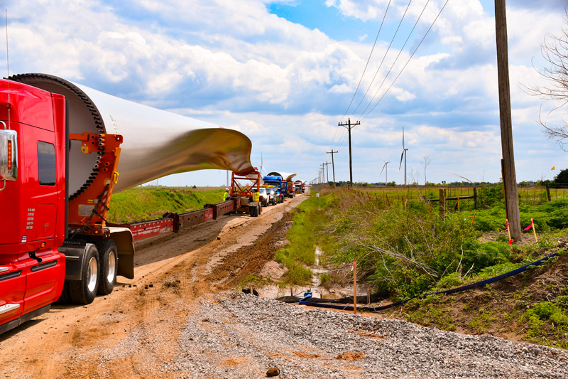 A red semi-truck transports a large wind turbine blade along a rural dirt road, with other vehicles and power lines visible, and wind turbines in the background under a partly cloudy sky.