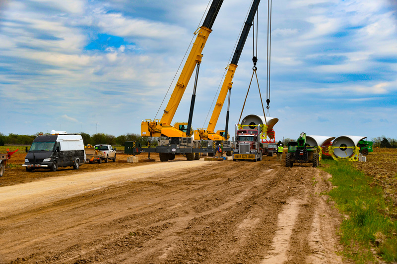 Two large cranes lift a wind turbine component onto a truck on a dirt road, surrounded by workers, vehicles, and construction equipment under a partly cloudy sky.