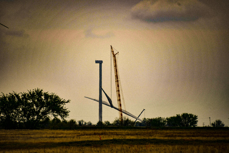 A large crane is helping to assemble or repair a wind turbine in a rural field with trees in the background under a cloudy sky.