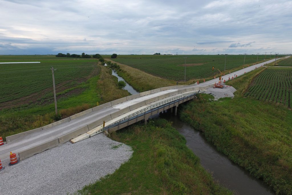 A rural road bridge under construction spans a small creek between green fields, with construction cones and barriers marking the work area. The sky is mostly cloudy, creating a calm atmosphere.