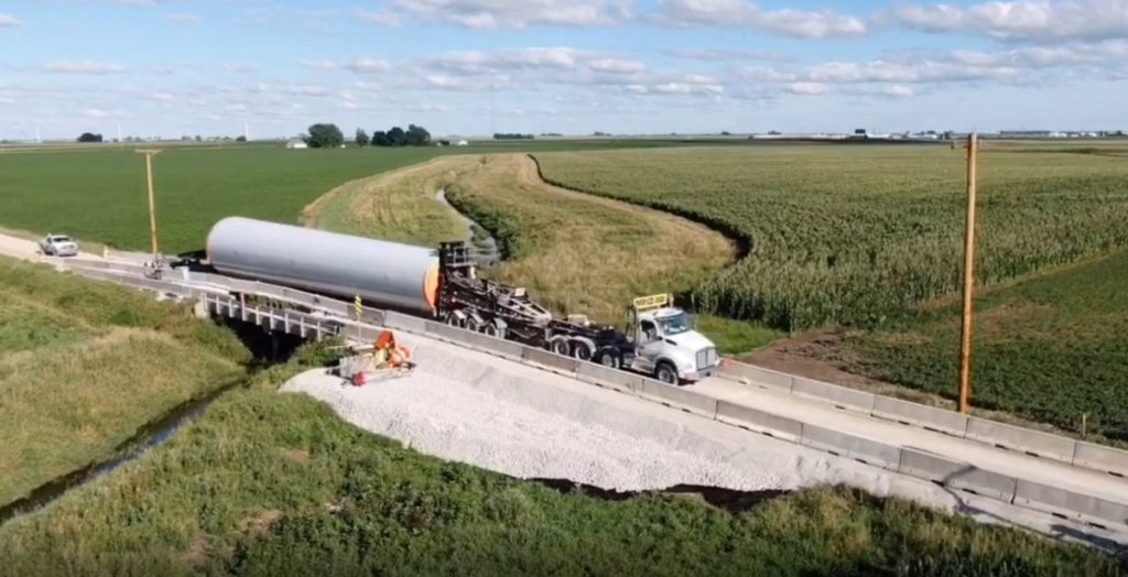 A large truck transports a long, cylindrical wind turbine blade on a rural road bridge surrounded by green farmland under a partly cloudy sky. Construction workers and gravel are visible near the bridge.