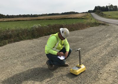A construction worker in a hard hat and safety vest kneels on a gravel road, writing on a clipboard next to a yellow soil compaction testing device, with fields and a road in the background.
