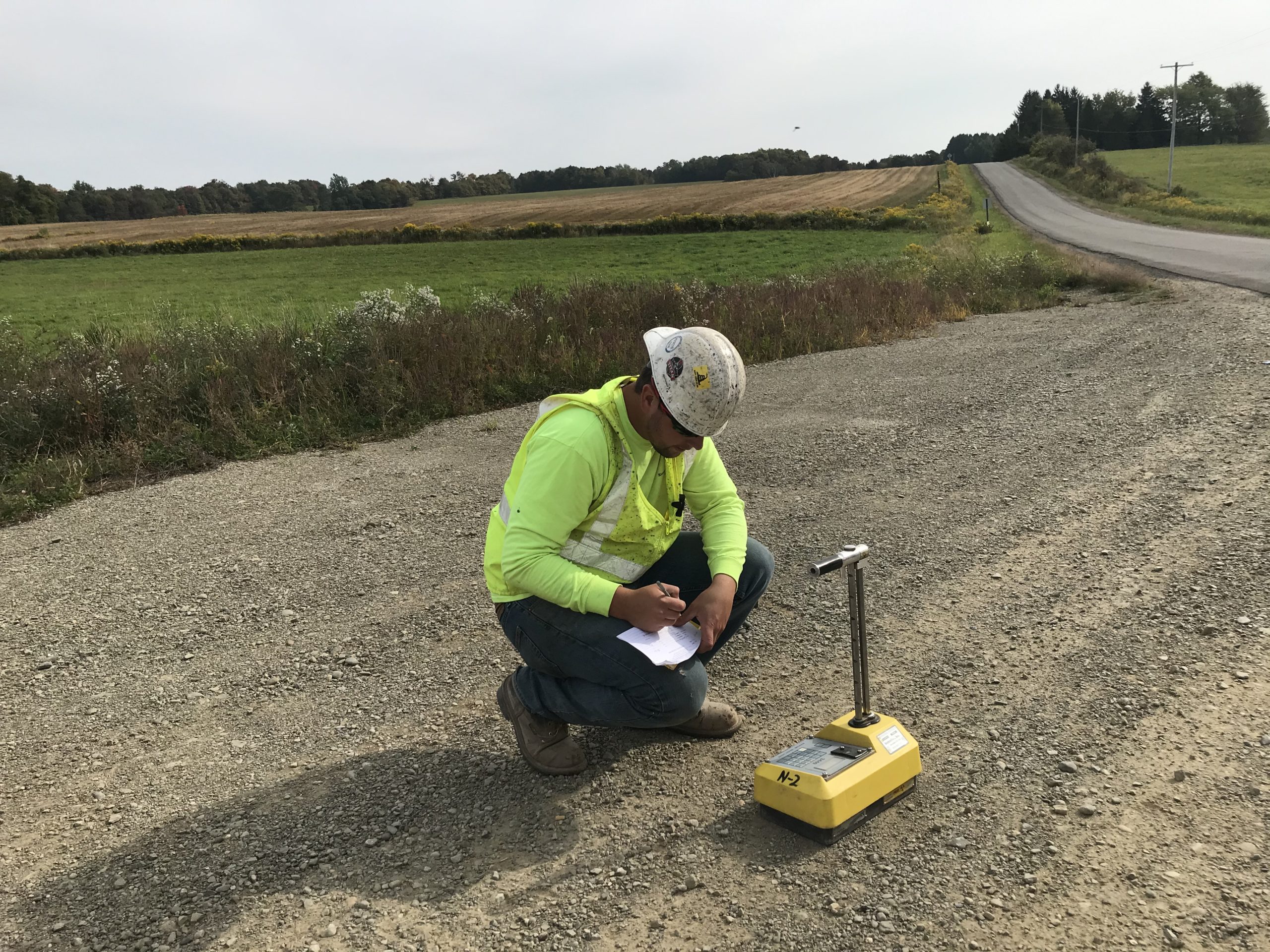 9-29 Post A construction worker in a hard hat and safety vest kneels on a gravel road, writing on a clipboard next to a yellow soil compaction testing device, with fields and a road in the background.