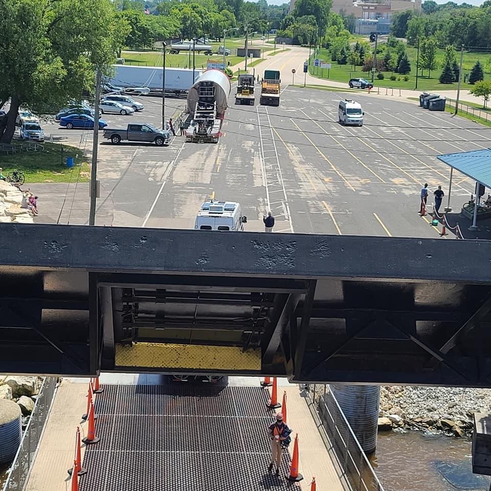 View from a ferry ramp showing a mostly empty parking lot, vehicles including a white van, a bus, and a pickup truck, with a bike rider exiting the ferry and people standing nearby on a sunny day.