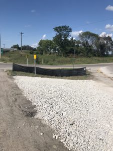 A gravel-covered area beside a road, bordered by a low black silt fence with yellow marker posts. Green grass and trees are visible in the background under a blue sky with scattered clouds.
