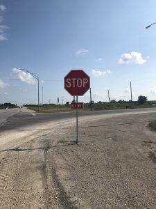 A stop sign with an All Way sign underneath stands at a rural intersection on a sunny day, with a paved road, gravel shoulder, grassy fields, and blue sky with a few clouds in the background.