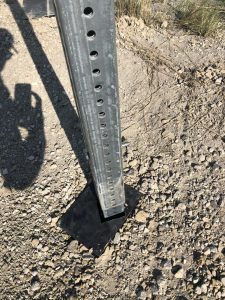A metal signpost is bolted to a black square base, set on rocky ground. A shadow of a person taking a photo is visible on the left side of the image. Sparse grass can be seen in the background.