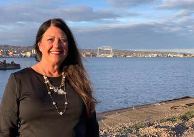 A woman with long dark hair, wearing a black top and necklace, stands smiling near a waterfront with a cityscape and cloudy sky in the background.