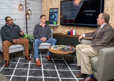 Three men sit in a modern office, engaged in conversation. Two wear casual business attire, one in a suit. A TV displays 2020 Roaring Twenties Logistics Group. Magazines and decor are on a round table on a patterned rug.